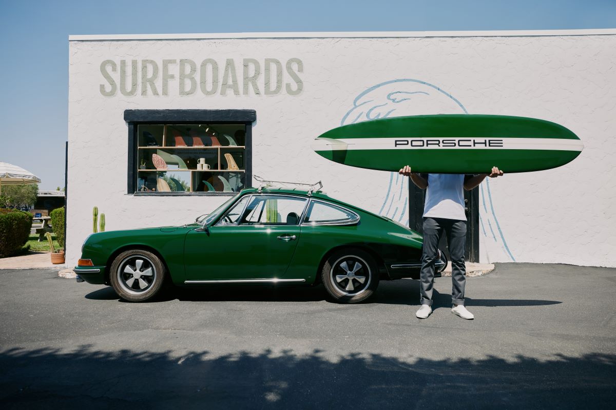 A man standing next to a vintage green Porsche with a surfboard reading, "Porsche"